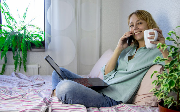 年轻女子躺在卧室的床上，在笔记本电脑上工作，在家里打电话。在家工作的概念，通过互联网远程在线工作。图片下载