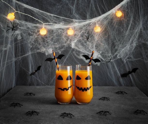 万圣节南瓜或胡萝卜饮料的杯子与南瓜灯的脸。在深灰色的背景上用果汁、发光的花环和蜘蛛网装饰万圣节派对。图片下载