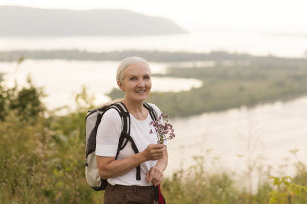 照片中，一位面带微笑的老年妇女身穿一件白色t恤，手里拿着一束草药，背着背包，悠闲地望着相机。副本的空间。图片下载