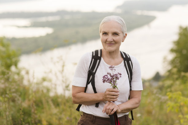 照片中，一位面带微笑的老年妇女身穿一件白色t恤，手里拿着一束草药，背着背包，悠闲地望着相机。副本的空间。图片下载