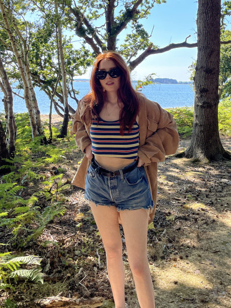 女人的形象，长，红色的头发拉牛仔短裤腰带在腰部，穿着夹克，条纹背心顶部和镜像太阳镜，站在海岸线林地阴影，看着相机，蓝色的夏天的天空，大海，重点在前景图片下载