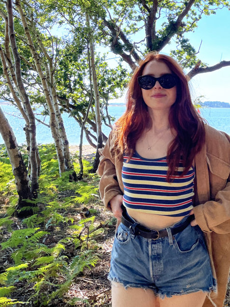 特写形象的女人长，红色头发拉牛仔短裤腰带在腰部，穿着夹克，条纹背心顶部和镜像太阳镜，站在海岸线林地阴影，看着相机，蓝色的夏天的天空，大海，重点在前景图片下载