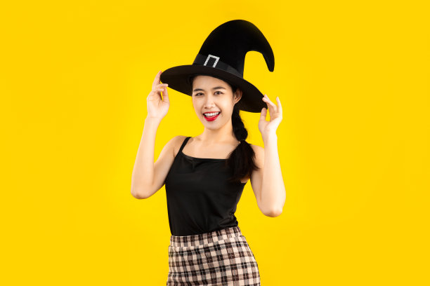 年轻的亚洲女子在黑色背心，戴着女巫帽在黄色背景为万圣节的概念。图片下载