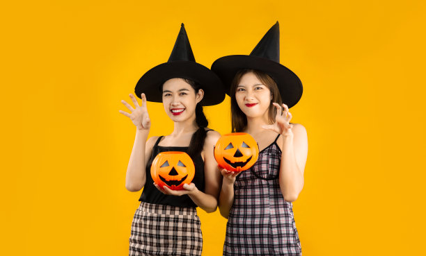 万圣节前夕，年轻的亚洲女孩穿着黑色连衣裙，戴着女巫帽，拿着南瓜桶在黄色背景上摆出不给糖就捣蛋的姿势。图片下载