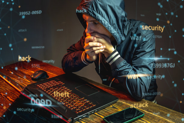 戴着兜帽、拿着手机的黑客正在一间黑暗的房间里用笔记本电脑键盘打字。网络战争和Dos攻击的概念图片下载