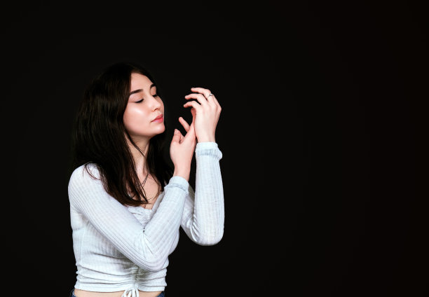 美丽的年轻女子，黑色长发，白色衬衫，闭着眼睛，轻轻地搓手。隔离在黑色背景。概念化妆品，皮肤治疗，护肤与copyspace。图片下载