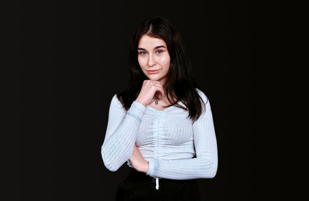 身材苗条的年轻女子身穿白色衬衫，胸部丰满，用手托着头，在黑色背景下对着镜头摆姿势，看上去若有所思。横幅，放置文本的地方。图片下载