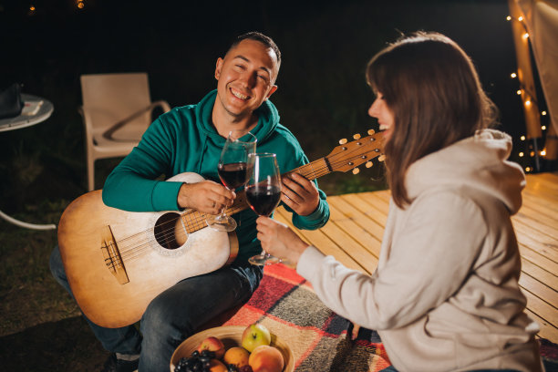 秋天的晚上，一对幸福的夫妇在豪华野营中放松，在舒适的篝火旁喝着酒，弹着吉他。户外休闲休闲的豪华露营帐篷。生活方式的概念图片下载