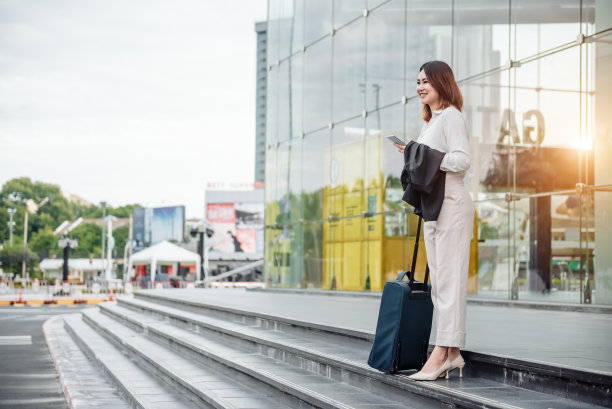亚洲妇女在登机口用智能手机在候机楼等候。空中旅行的概念与年轻休闲的商务女性坐着聊天的智能手机，随身携带的手提行李推车。图片下载
