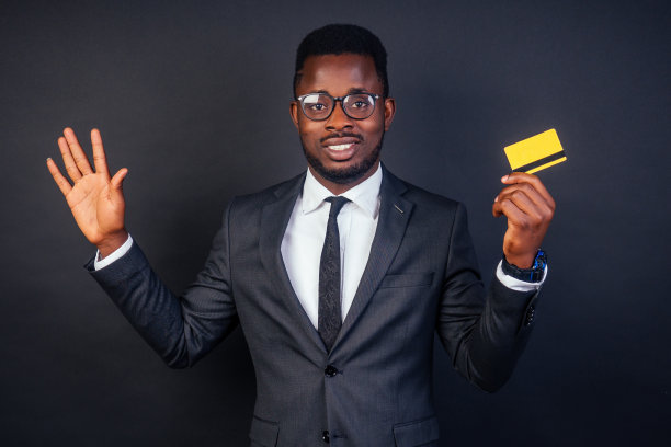 年轻的成功商人穿着时尚的黑色经典套装，戴着酷炫的眼镜，拿着一张黄色塑料信用卡在工作室的深色背景上。购物的概念图片下载