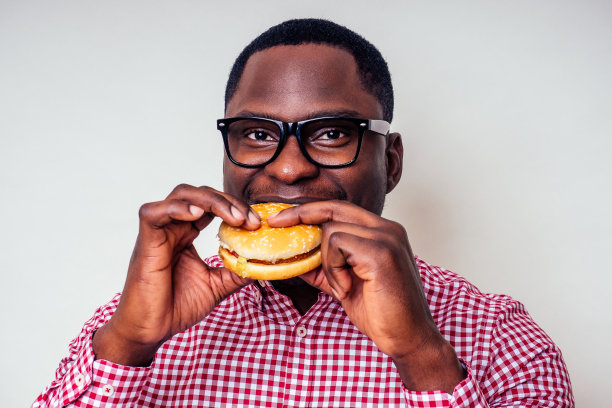 非裔美国人正在享受汉堡包的味道。英俊年轻的非洲裔男子，穿着时髦的衬衫，戴着眼镜，在白色背景下拿着一个汉堡。爱吃垃圾食品减肥图片下载