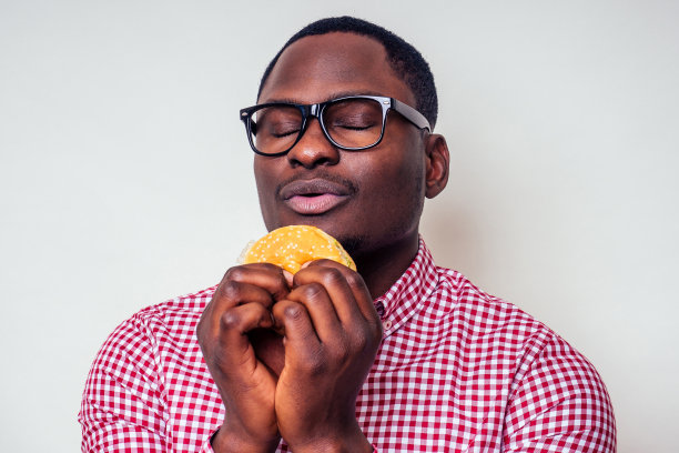 非裔美国人正在享受汉堡包的味道。英俊年轻的非洲裔男子，穿着时髦的衬衫，戴着眼镜，在白色背景下拿着一个汉堡。爱吃垃圾食品减肥图片下载