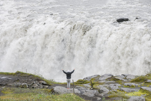 高角后视图年轻男子高举手臂站在一个强大的瀑布顶部图片下载