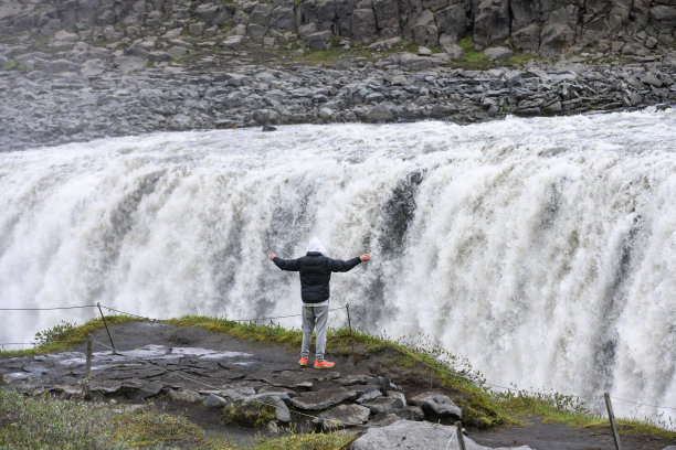 后视图，年轻男子高举双臂站在一个强大的瀑布顶部图片下载