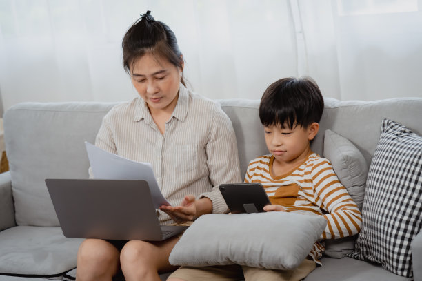 英俊的亚洲男孩，他的妈妈正在用笔记本电脑，小男孩很生气，紧张地从妈妈手里把纸拿过来，想让她自己玩。图片下载