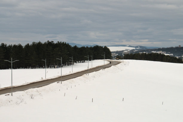冬天的景色:弯弯曲曲的山路，路灯，雪原和森林。天空乌云密布，远处城镇清晰可见图片下载
