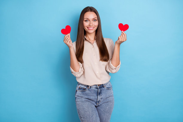 照片甜美漂亮可爱快乐的女士穿着时髦的衣服，举起手臂，展示红色的纸符号心脏孤立在蓝色的背景上图片下载