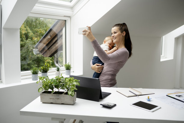 漂亮的年轻女子，女商人站在办公桌前，抱着睡着的儿子在她的怀里，在笔记本电脑，智能手机上工作图片下载