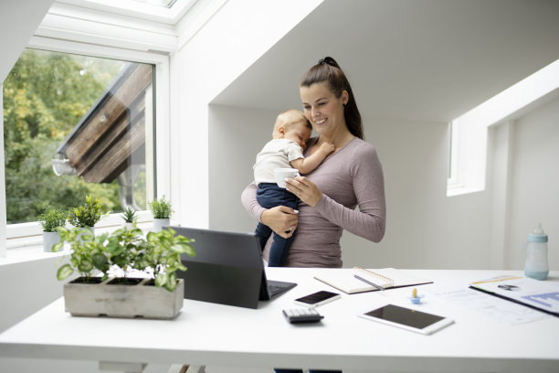 漂亮的年轻女子，女商人站在办公桌前，抱着睡着的儿子在她的怀里，在笔记本电脑，智能手机上工作图片下载