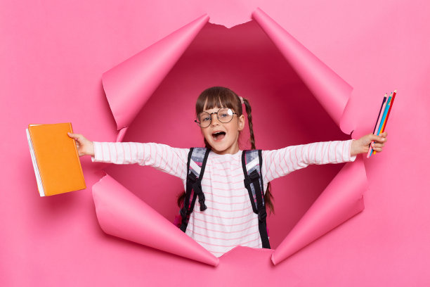 室内拍摄的快乐积极的小女生穿着衬衫和眼镜，背着粉色纸撕成碎片的背包站在那里，拿着铅笔和书，举起胳膊，高兴的样子。图片下载