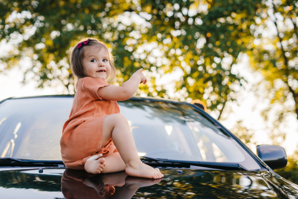 一个小女孩在她父母的汽车引擎盖上放松的画像，在他们的旅行途中，她的车停在路边。暑假。阳光明媚的一天。幸福的家庭,童年。儿童保健。图片下载