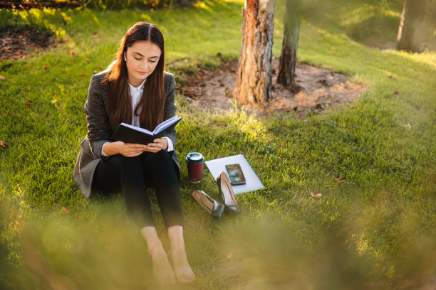 微笑着美丽的黑发女人坐在绿色的草地上读书和工作在公园里的夏天。教育和创造力。女学生在大学学习后放松图片下载