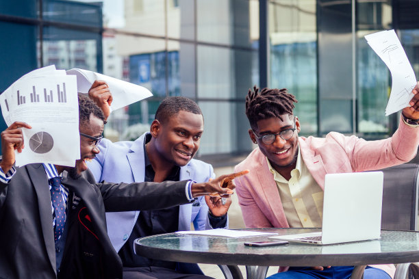 他们的三个成功的非裔美国商人穿着时髦的西装坐在桌子旁，用笔记本电脑进行在线会议，并把文件扔了起来。团队合作与成功理念图片下载