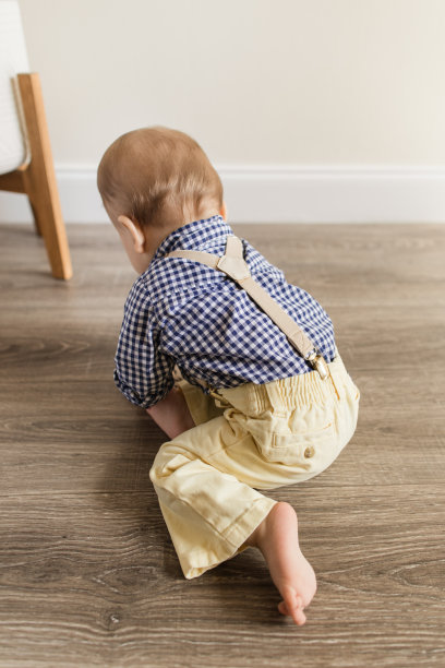 一个有12个脚趾的9个月大的大流行男婴，穿着海军蓝和白色的格子衬衫，带背带和浅黄色的裤子，在地板上玩耍图片下载