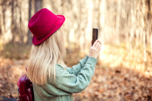 背影上一个女孩戴着红帽子，背着背包拿着手机，拍摄着美妙的秋景。图片下载