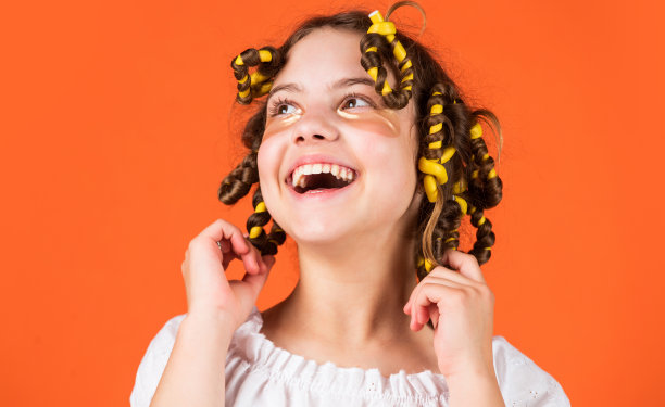 可爱的儿童发型。戴着卷发器的女儿在笑。造型技巧。青少年的爱好。小女孩卷发使用卷发器橙色背景。理发师沙龙。女性美程图片下载