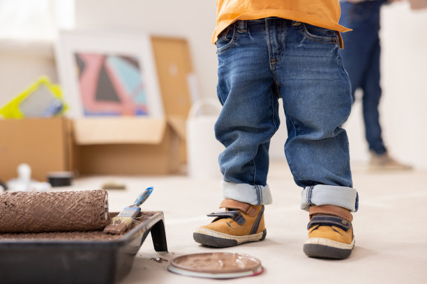 一个孩子的腿特写，穿着牛仔裤和橙色运动鞋。侧面是一个装有油漆的等离子碗和油漆辊。背景是需要修理的东西图片下载