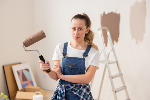 一个年轻的女学生站在她的宿舍正在装修的房间中间。这是她的第一次经历。她右手拿着一个油漆滚筒图片下载