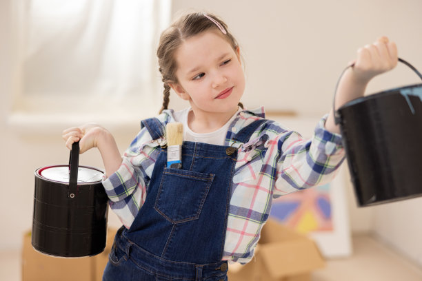 女孩穿着牛仔工装裤，白色t恤和格子衬衫，双手拿着黑色油漆罐。孩子饶有兴趣地审视着它们，想象着未来孩子的房间会是多么美丽图片下载