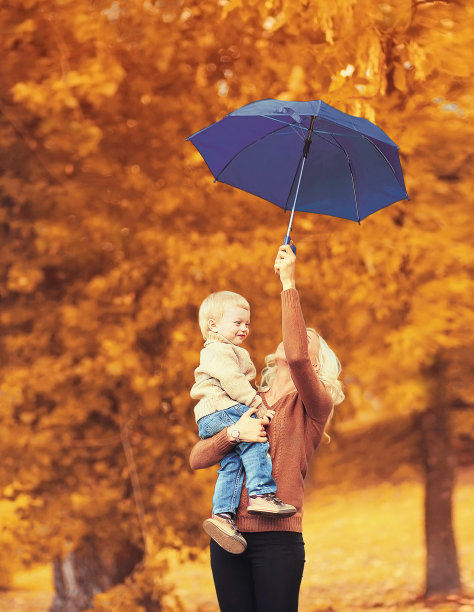 在秋天的树背景下，快乐微笑的母亲抱着打着伞的孩子一起玩耍图片下载
