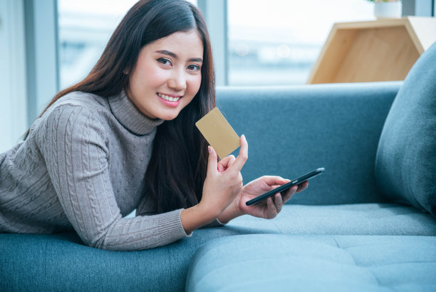 女人坐在沙发上用智能手机，用信用卡在网上购物，带着笑容的购物狂人快乐的时光。快乐的亚洲女人躺在智能手机旁。女性网购支付素材图片