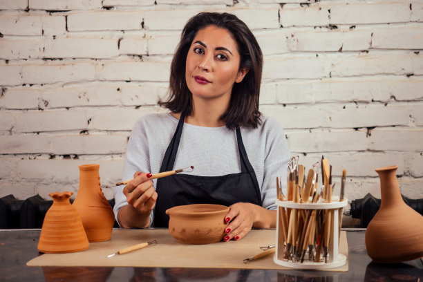 年轻迷人的女陶工在工作室工作，手拿工具和杯子在工艺车间的棕色陶瓶壶。陶器店的女商人图片下载