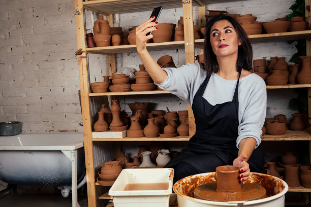 商业女性陶工自拍照片在智能手机上的自画像作品与粘土陶轮在一个创意工作室的艺术雕塑家学院的学生图片下载