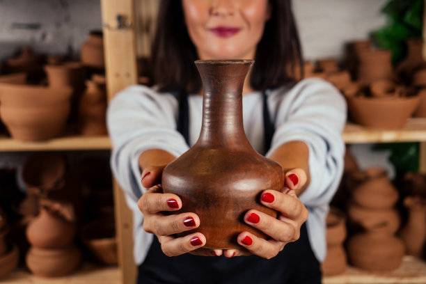 年轻迷人的女陶工在工作室工作，手拿工具和杯子在工艺车间的棕色陶瓶壶。陶器店的女商人图片下载