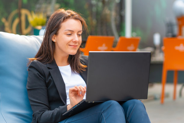 年轻微笑的女商人在笔记本电脑键盘上打字。一名女性在户外咖啡馆用笔记本电脑工作。夏天有风的日子。图片下载
