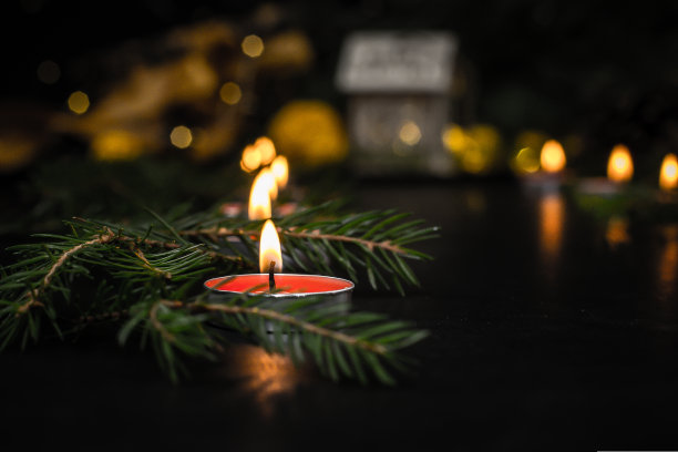 在一张黑色的桌子上，一根大蜡烛正在燃烧，近处是一棵圣诞树的树枝，上面散落着绿色的小针。模糊的背景显示圣诞彩灯和玩具房子。图片下载