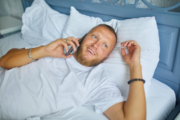 大胡子男人早上在家里躺在舒适的白色亚麻布床上说着智能手机，男人在卧室里聊天，打电话打发时间图片下载