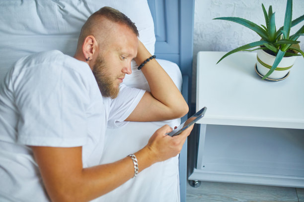 大胡子男子在早上使用智能手机在舒适的床上，而躺在家里的白色亚麻布，男性聊天，上网上网花时间在卧室图片下载