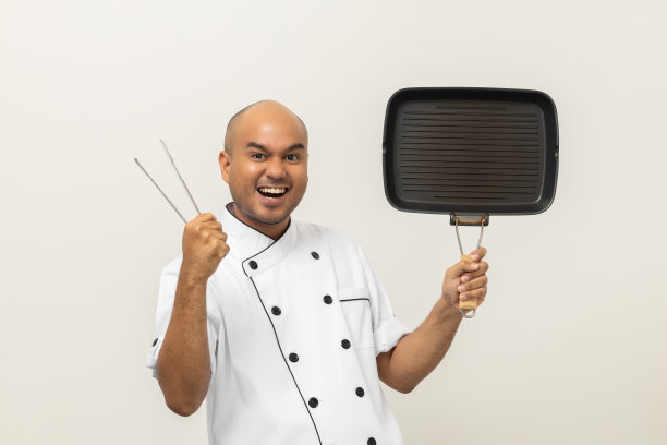 年轻的亚洲男性厨师在统一烹饪铁制烧烤锅和钳子厨房用具。厨师职业厨师在厨房、餐厅和酒店工作的人。图片下载