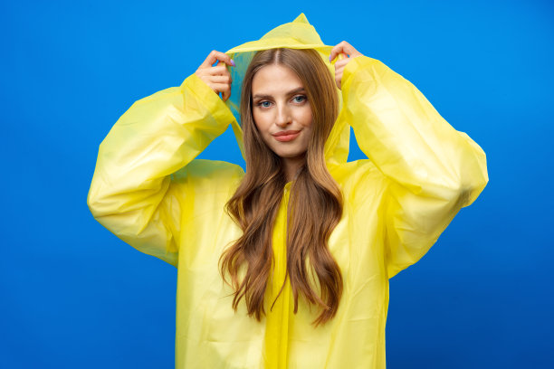 年轻的金发女人穿着黄色雨衣微笑的镜头在蓝色的背景在工作室图片下载