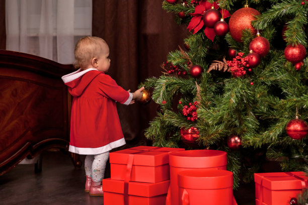 后视图在室内装饰的圣诞树背景下，一个一岁的小女孩伪装成圣诞老人。婴儿在圣诞节的家庭装饰。温馨家的新年庆祝概念。复制空间，文本位置图片下载