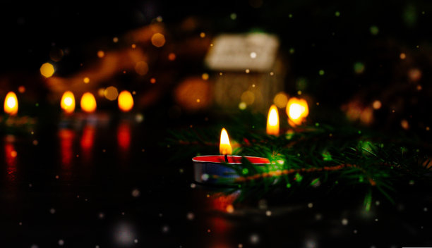 圣诞之夜，黑色背景上燃着红色蜡烛的火焰，附近躺着一棵圣诞树的树枝。在背景中，一个模糊的背景勾勒出一个玩具木屋。图片下载
