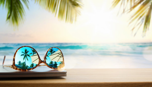 电脑和太阳镜放在木桌上。在落日的背景下，温暖的热带海滩上的太阳图片下载