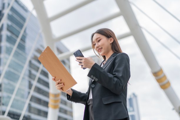 迷人的年轻亚洲职业女商人女士与一个美好的微笑夹夹板看着电话检查市场和销售库存报告在曼谷市的中央商务区图片下载