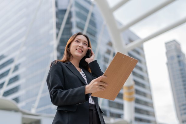 在曼谷市中心的一个商业区内，年轻迷人的亚洲职业女商人女士面带微笑，在看完剪贴板上最新的市场和销售报告后，抬头看向天空，一边说着电话图片下载