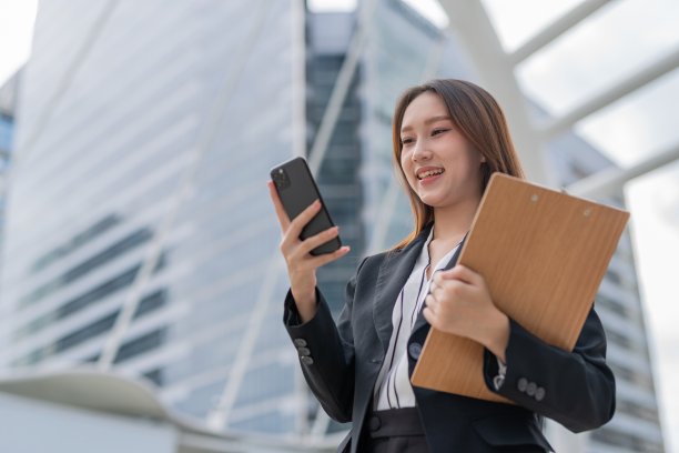 迷人的年轻亚洲职业女商人女士，微笑着，拿着夹夹板，看着电话，检查会议和销售库存报告在曼谷市的中央商务区图片下载